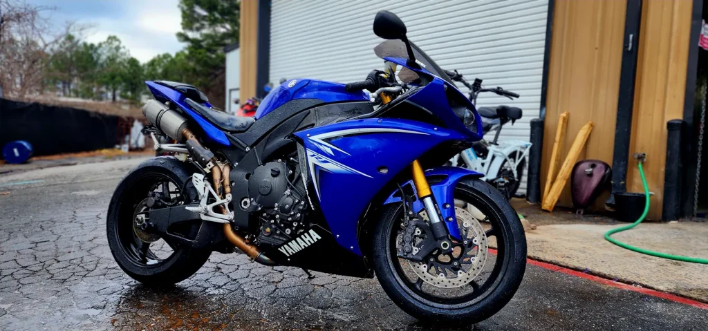 Clean blue Yamaha YZF-R1 with gold forks parked on wet pavement outside the shop bay, full side profile.