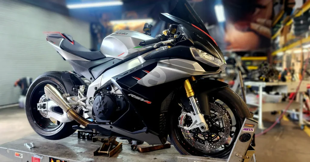 Clean silver and black Aprilia RSV4 on a lift under shop lighting, glossy bodywork and polished exhaust, no work in progress.