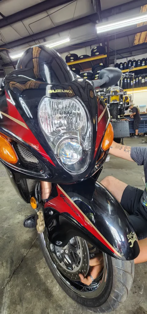 Close-up front three-quarter of a black and red Suzuki Hayabusa on the shop floor with a small gold Diaz logo decal on the front fender and a person's legs beside it.