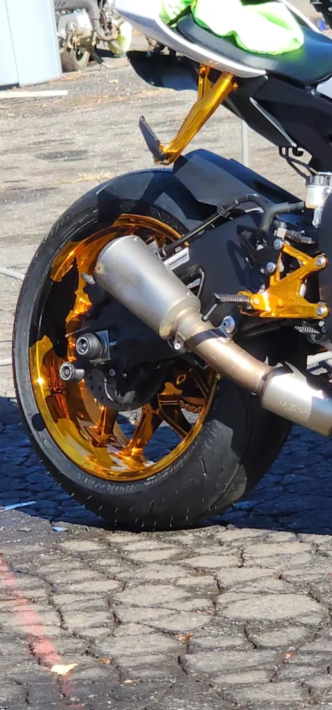 Close-up of a sportbike rear wheel with gold anodized wheel, gold rearsets and a titanium slip-on exhaust, shot on pavement.
