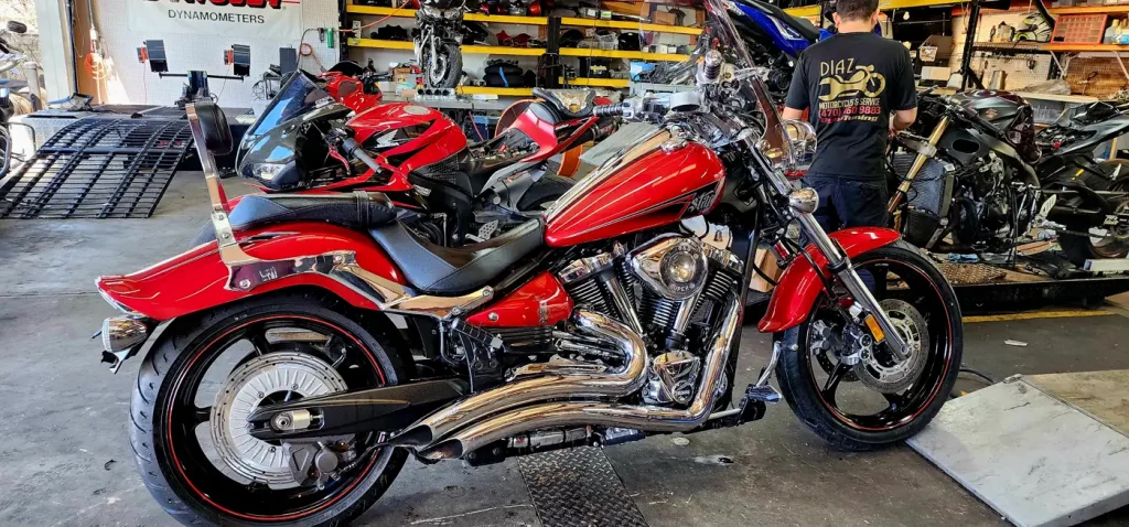 Polished red Yamaha Raider/Warrior cruiser with heavy chrome parked on the shop floor while a worker in a Diaz shirt stands behind it.