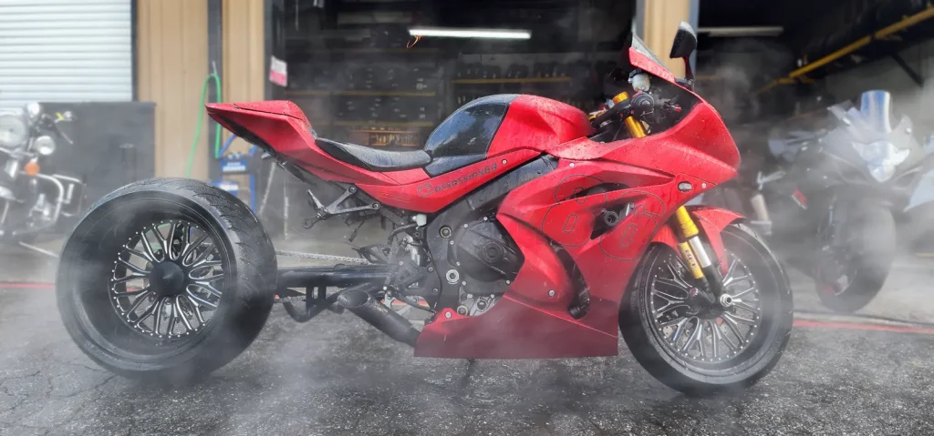 Red fat-tire Suzuki GSX-R custom being rinsed outside the shop bay, heavy water mist surrounding the bike.