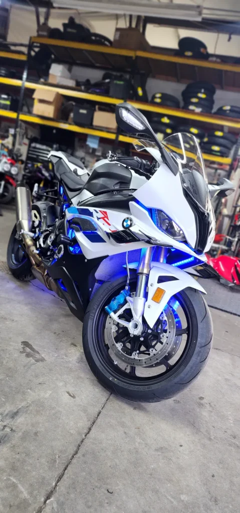 White, blue and red BMW S1000RR with blue LED underglow lit up on the shop floor, tire racks behind, shot from a low front three-quarter angle.