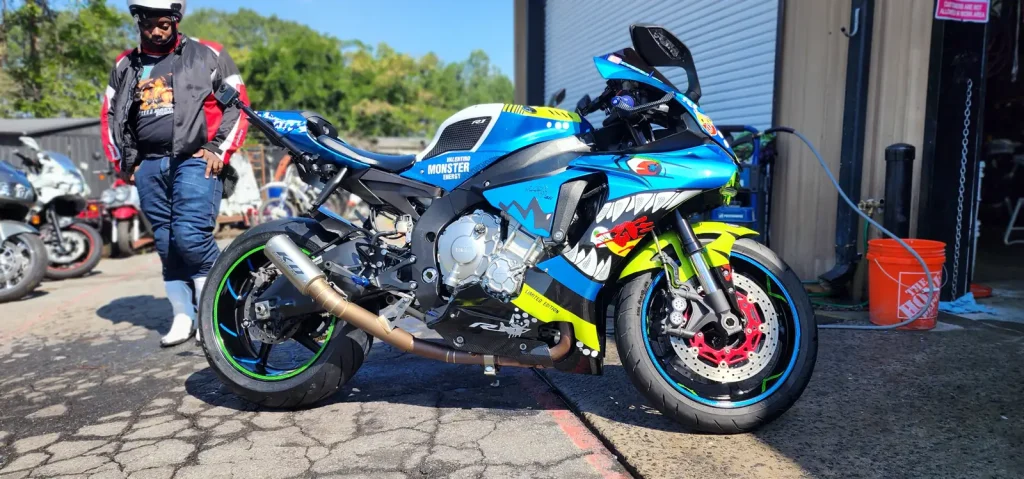 Yamaha R1 in Valentino Rossi shark-face livery being washed outside the bay with a hose and orange bucket, a rider in a jacket standing nearby