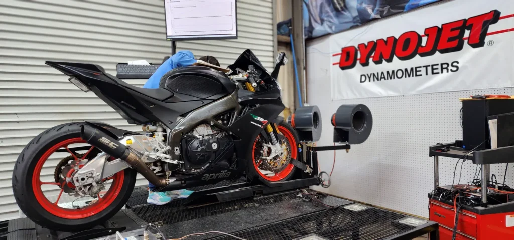 Black Aprilia RSV4 strapped down on the Dynojet dyno with the tail sections removed and a technician leaning over the tank; large DYNOJET banner behind.