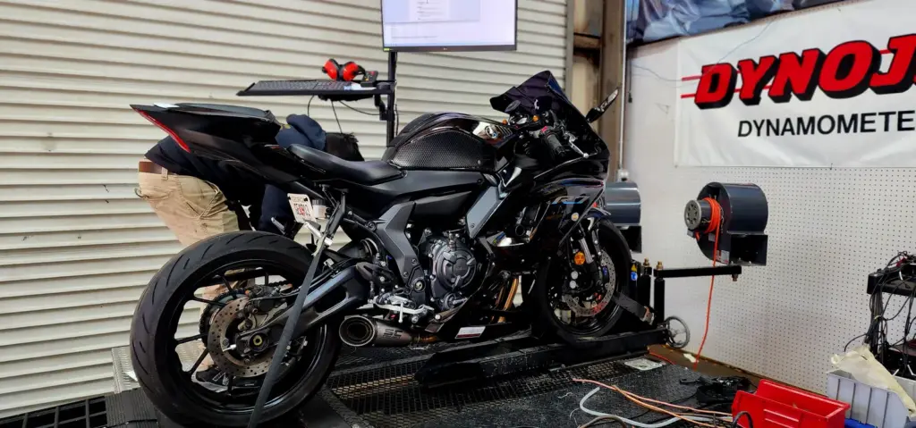 A black sport motorcycle sits on a dynamometer test bench with equipment and monitor in a garage setting.