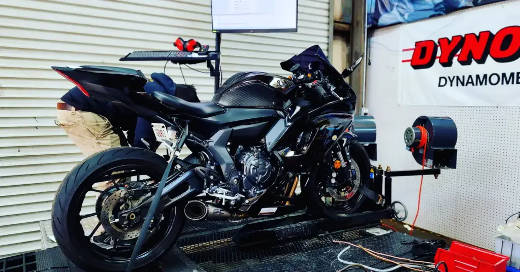 A black sport motorcycle sits on a dynamometer in a garage, surrounded by cables, equipment, and a monitor.