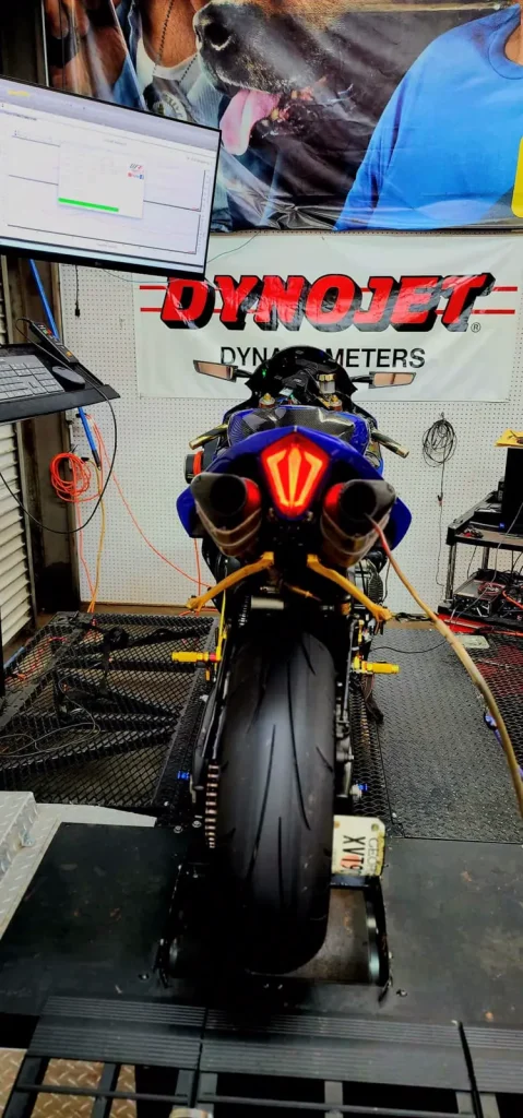 Rear view of a blue Yamaha R1 strapped on the Dynojet dyno drum with the tuning software graph on the monitor and a wideband probe in the exhaust.