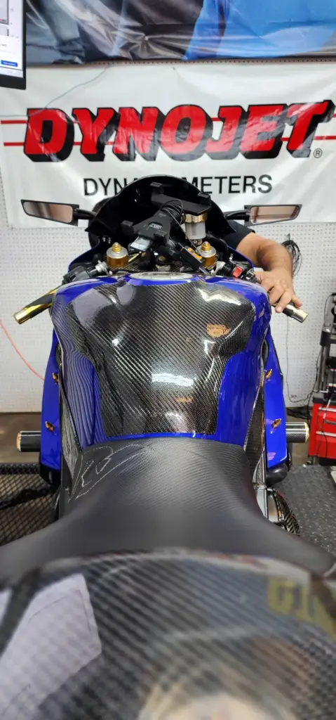A blue motorcycle with a carbon fiber tank sits on a dynamometer as an operator adjusts the controls.