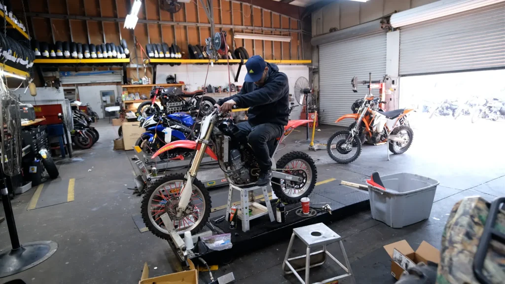 Technician sits on a red Honda dirt bike strapped down on a dyno platform inside the shop, with a stool under the bike and other bikes parked around.