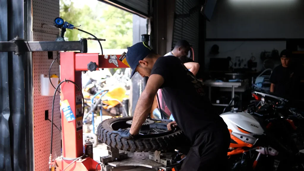 Technician bends over a knobby tire mounted on the red tire machine at the open bay door, with a white/orange KTM and other staff in the background.