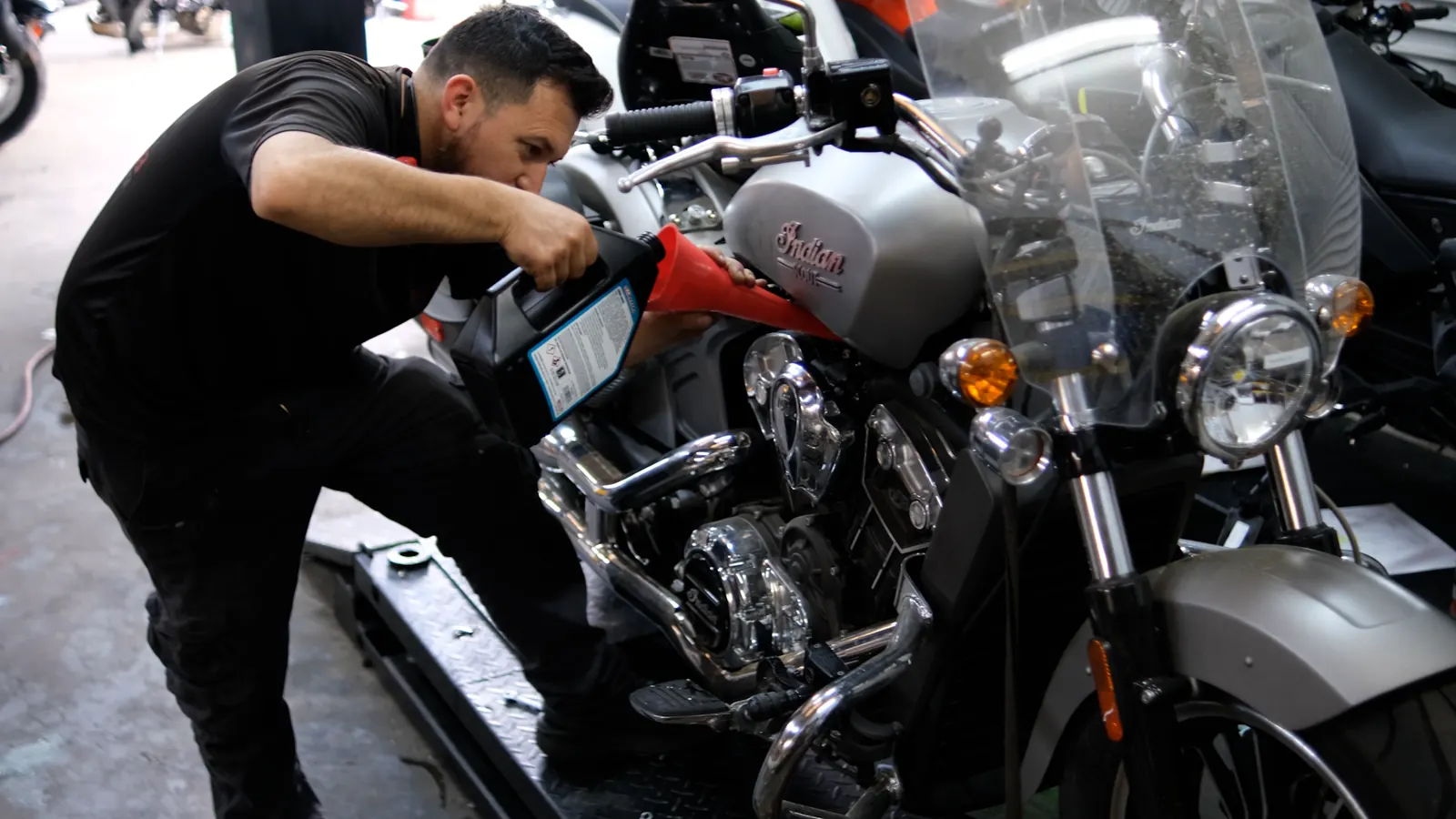 East Cobb motorcycle repair shop work — technician pouring oil through a red funnel into a silver Indian Scout on the lift