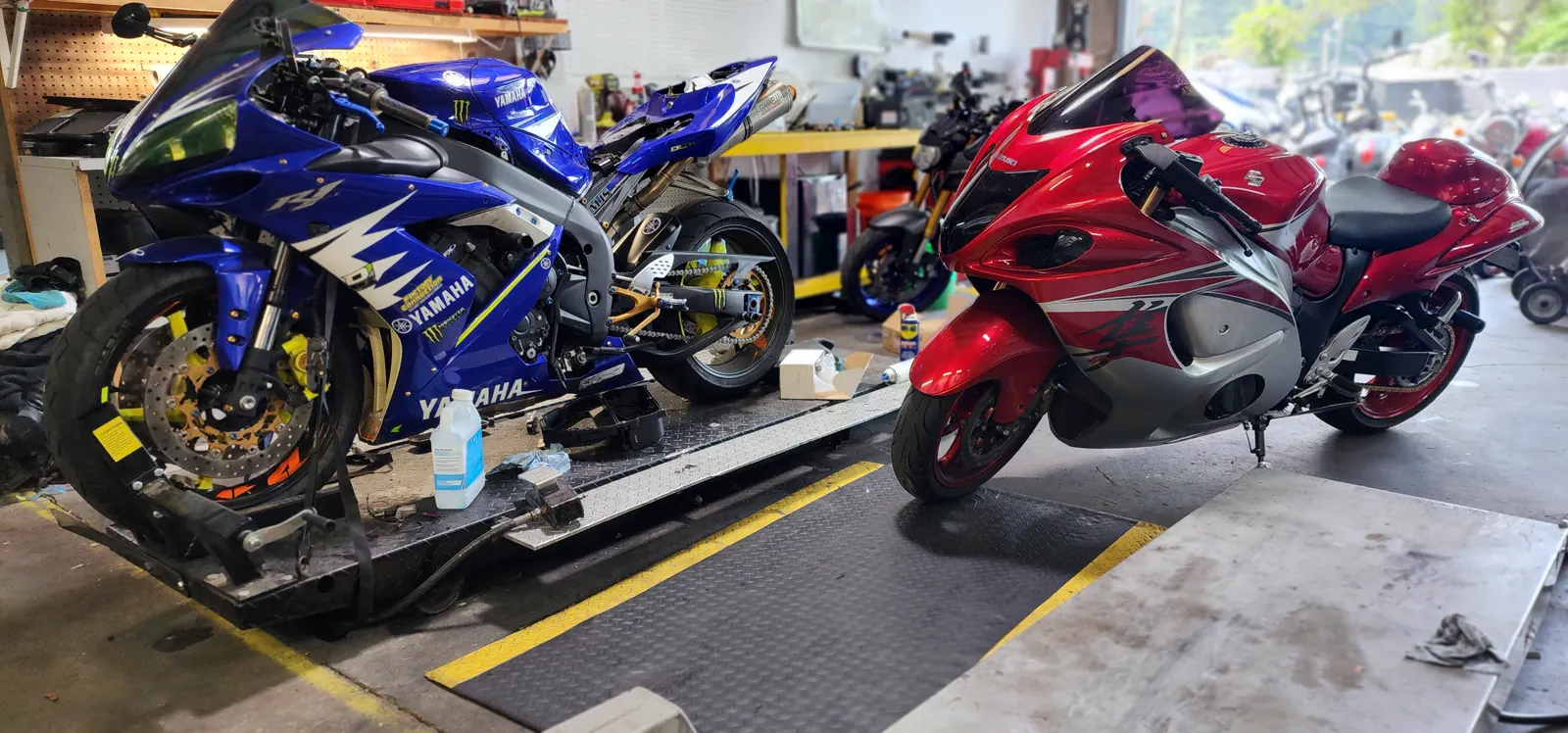Overdue motorcycle service Cartersville — blue Yamaha R1 on a lift beside a red Suzuki Hayabusa, fluid bottles nearby