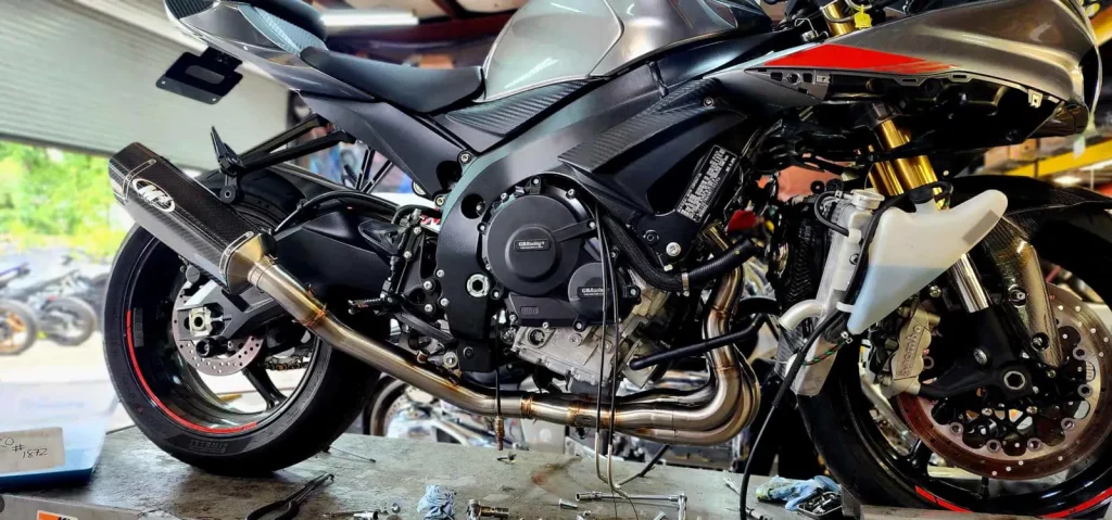 Close side view of a silver Suzuki GSX-R with fairings partially removed, showing engine cases, headers and an aftermarket M4 exhaust, tools scattered below.