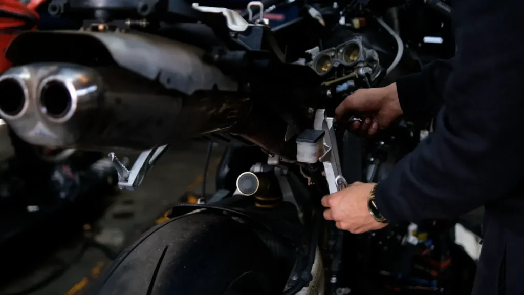 Close-up of hands using a wrench on the rear brake master cylinder / rearset area of a sportbike with a dual-outlet exhaust can visible at left.