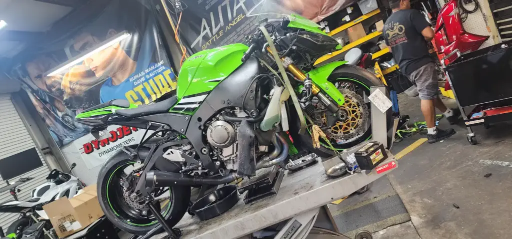 A green sport motorcycle is being repaired in a cluttered workshop with scattered tools and bike parts.