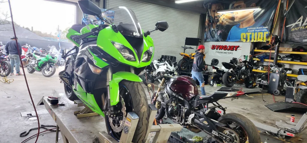 Green Kawasaki ZX-6R on a lift in the foreground while a stripped maroon bike sits mid-teardown and a tech in a red cap works near the Dynojet banner.