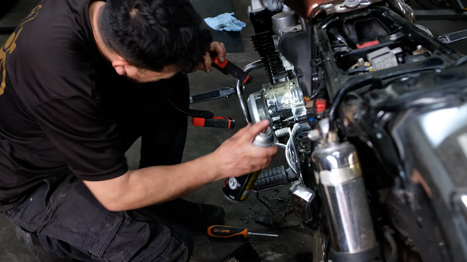 Motorcycle service Gainesville — kneeling tech spraying cleaner onto the chrome engine and carb area of a cruiser