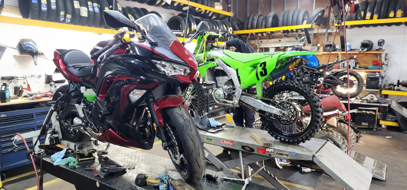 Motorcycle service Gainesville — maroon Kawasaki Ninja on a lift while a tech works on a green Kawasaki KX dirt bike nearby