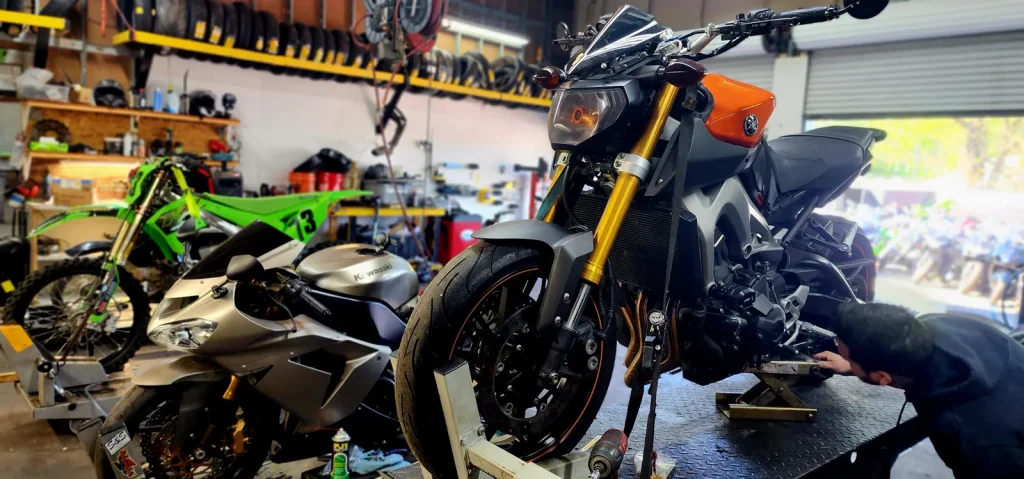 Orange and gray Yamaha FZ-09/MT-09 on a lift with a tech crouched at the right side working low on the bike; stripped Kawasaki and dirt bike behind.