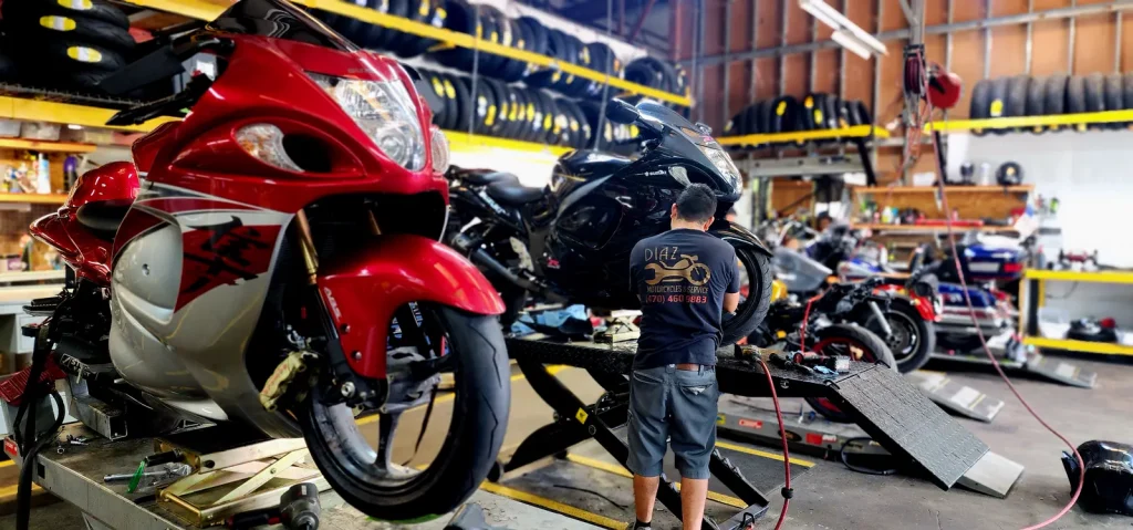 Red-and-silver Suzuki Hayabusa in the foreground on a lift while a tech in a Diaz shirt works on a black Hayabusa on a second lift, tire racks lining the walls.