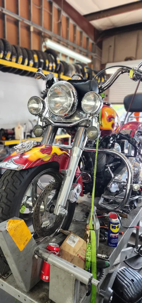 Red Harley-Davidson cruiser with orange flame paint sits strapped on a lift with tools, WD-40 and a parts box on the bench below; rows of tires hang overhead.
