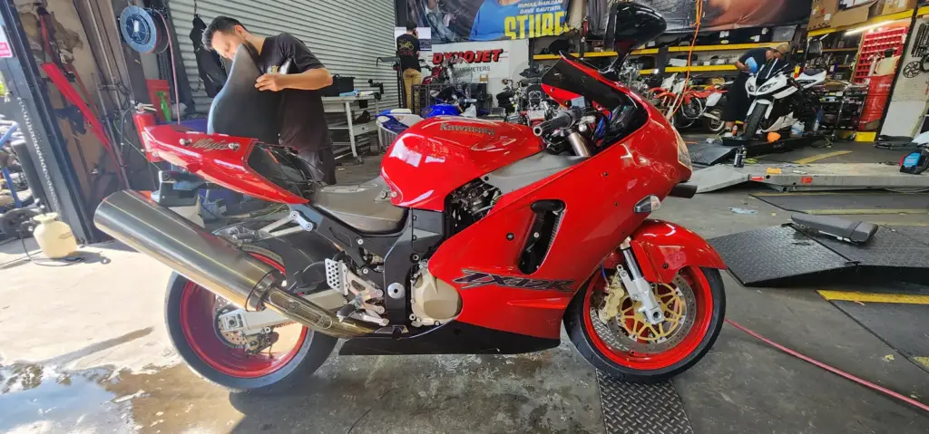 A shiny red Kawasaki Ninja motorcycle sits in a workshop while a person works in the background.