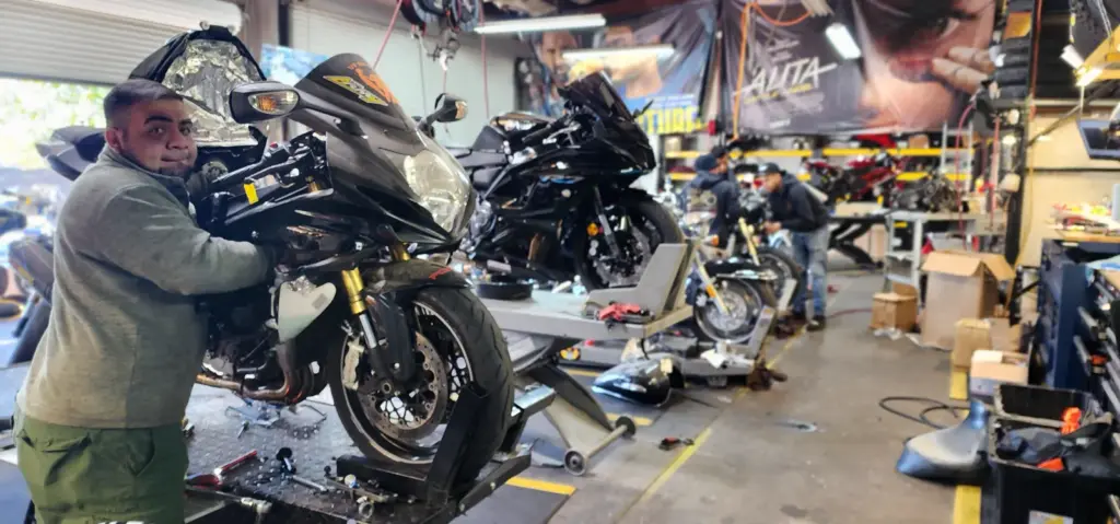 A mechanic repairs a black motorcycle inside a busy garage filled with bikes and various repair tools.
