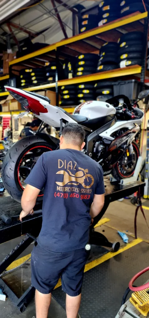 A tech in a Diaz Motorcycles shirt works at a lift beside a white Yamaha R1 with racks of tires stacked behind.