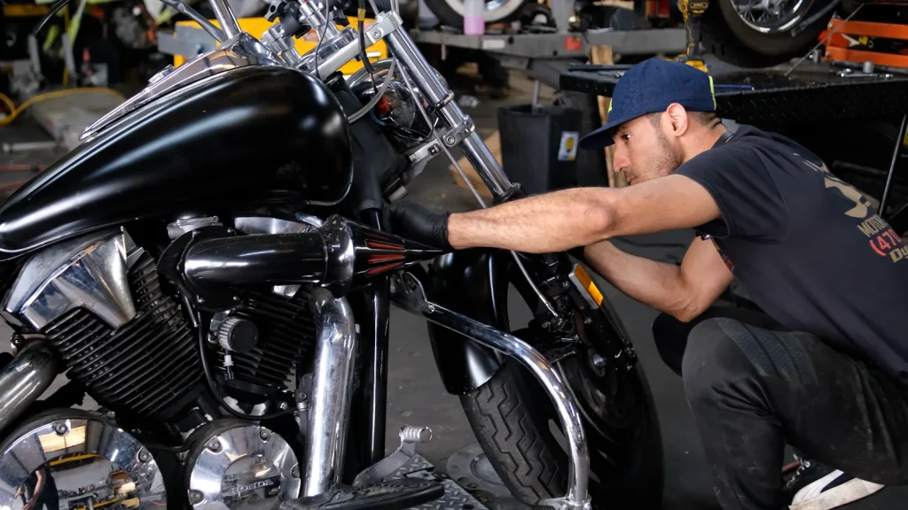 Technician in a cap and Diaz shirt crouched at the front wheel of a black Honda V-twin cruiser, hands on the brake/fork area.
