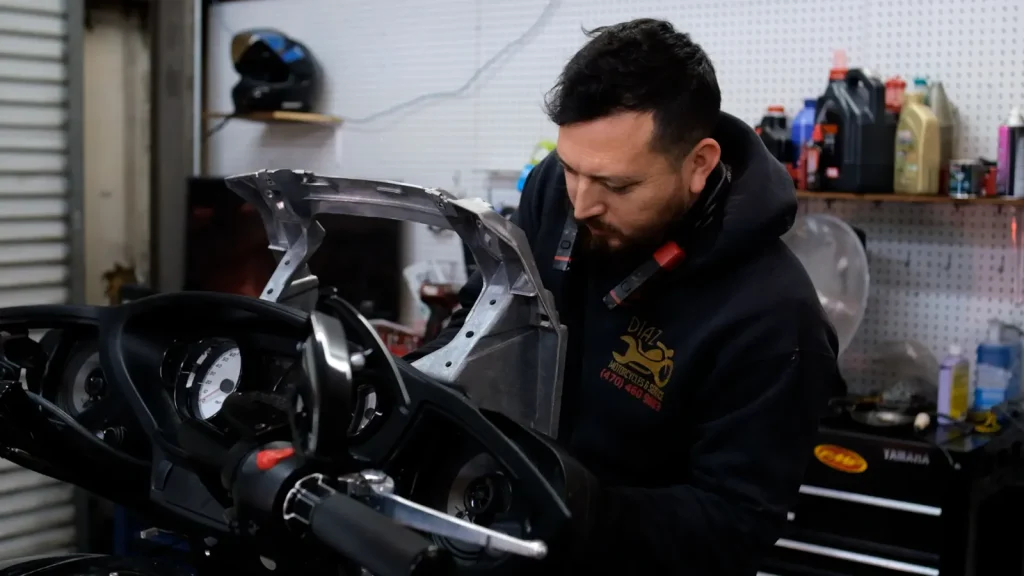 Technician in a Diaz Motorcycles hoodie removes or fits a fairing bracket/subframe above a touring bike's instrument cluster and handlebars inside the shop.