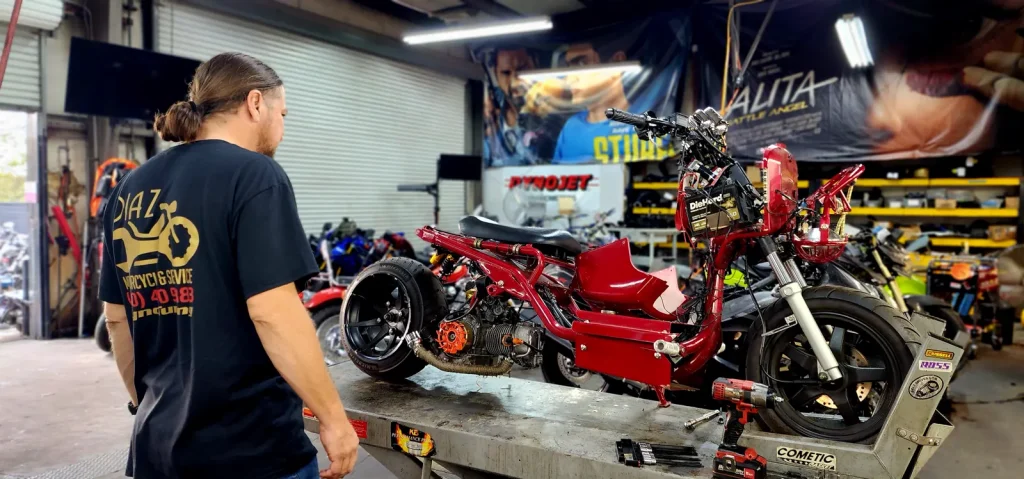 Technician in a Diaz Motorcycles shirt stands looking at a red custom Honda Ruckus on the lift table with side panels off and engine exposed.