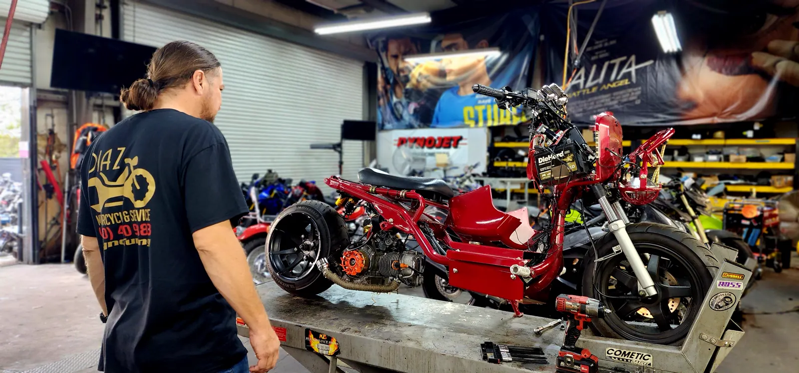 Motorcycle service near Kennesaw — technician in a Diaz shirt studying a red custom Honda Ruckus with side panels off