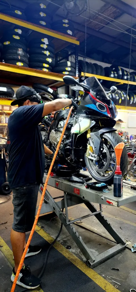 Technician leans over a BMW S1000RR with fairings removed on a lift table, oil drain pan and oil bottle on the deck.