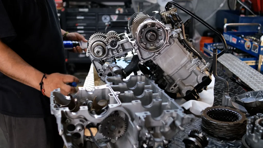 Technician spraying assembly lube from a blue can onto a disassembled engine and open crankcase on the bench.