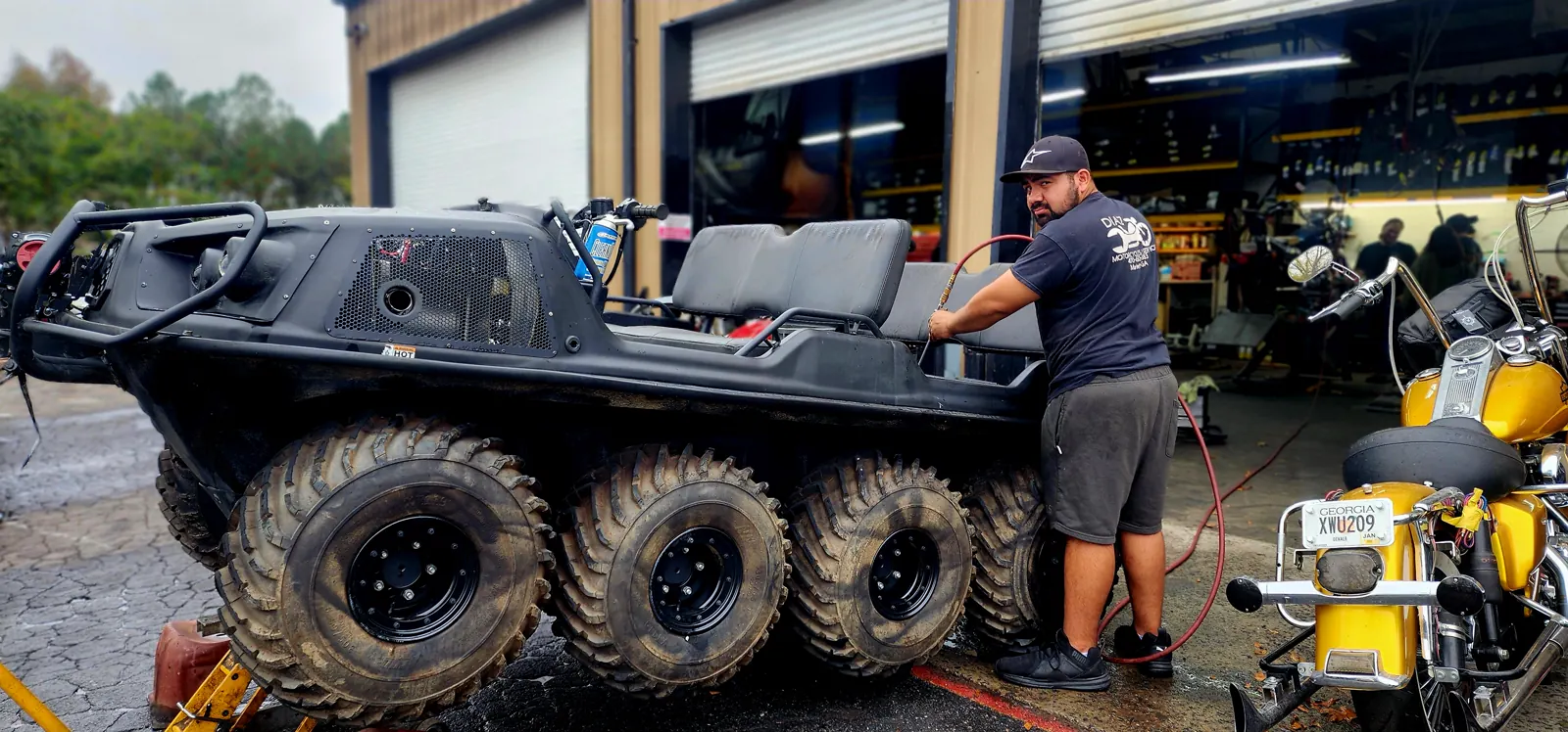 motorcycle service Newnan — technician with an air hose beside a black six-wheeled Argo ATV and a yellow Harley bagger
