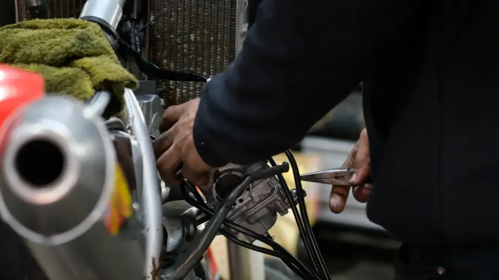 A person repairs a motorcycle engine using a wrench, with various tools and exposed wires visible nearby.
