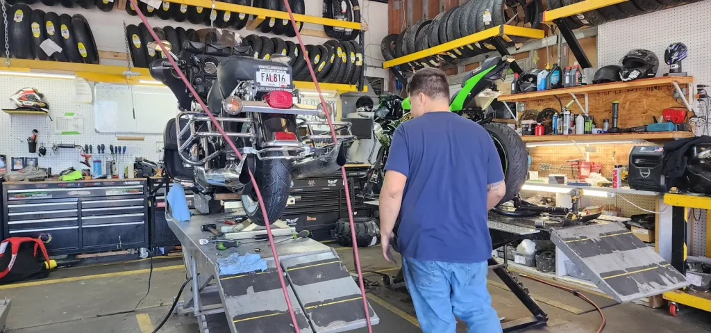Technician walks past a Harley touring bike raised high on a lift table with tools scattered on the bench, tire racks and pegboard filling the shop.