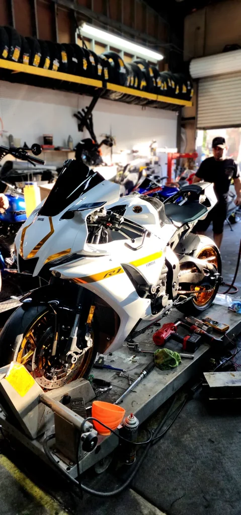 White Yamaha R1 with gold wheels and gold chain on a lift surrounded by tools, with a technician walking in the background.