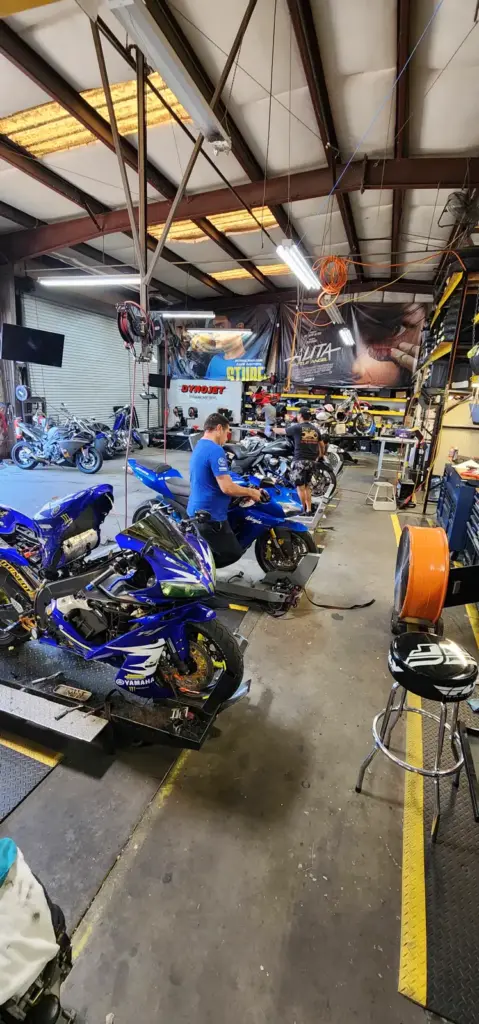 A mechanic repairs a blue motorcycle in a busy garage with bikes, tools, and equipment visible around.