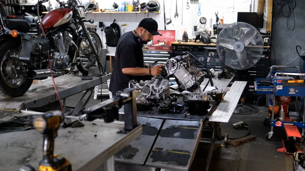 Wide shop shot of a technician at the bench working on a torn-down engine with parts spread across the table, maroon Honda Shadow on a lift at left and a shop fan behind.