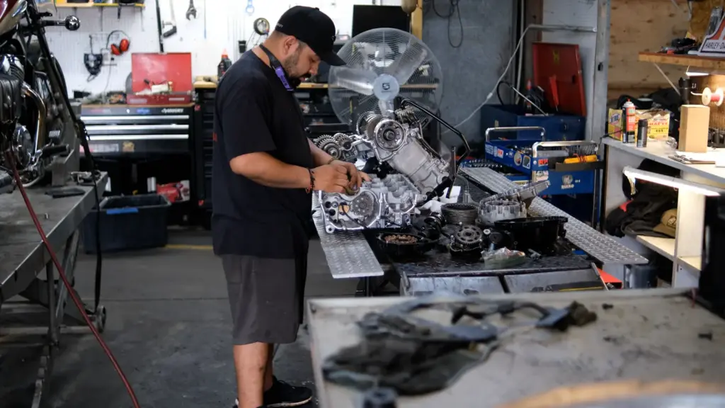 A mechanic in a black shirt and cap repairs a motorcycle engine in a cluttered workshop setting.