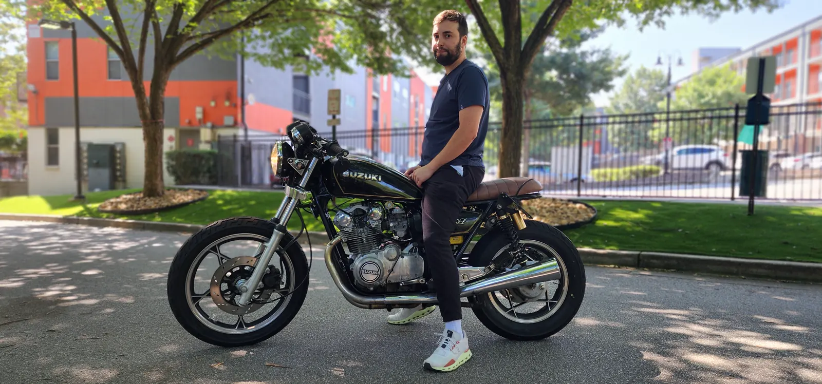 A man in dark clothes sits on a black Suzuki motorcycle parked in a shaded, urban outdoor setting.