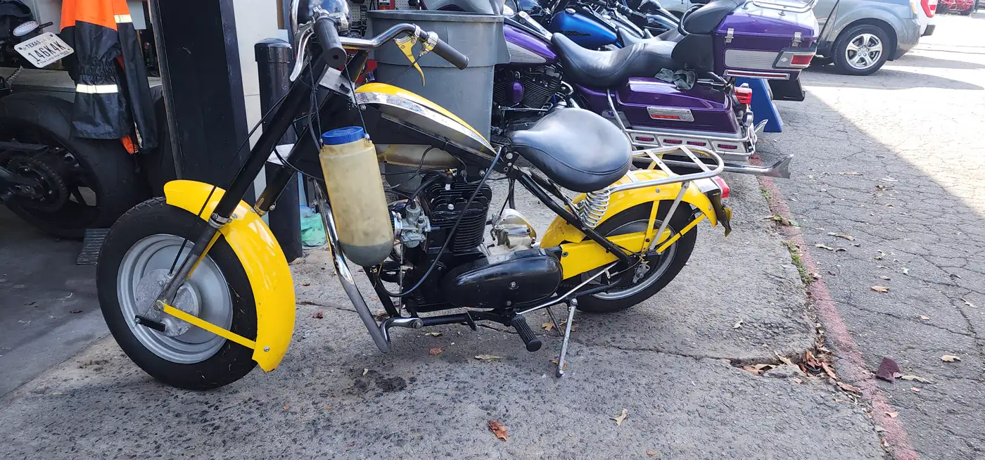 restore vs sell motorcycle Smyrna — vintage yellow and black Cushman-style scooter with springer front end on the shop apron