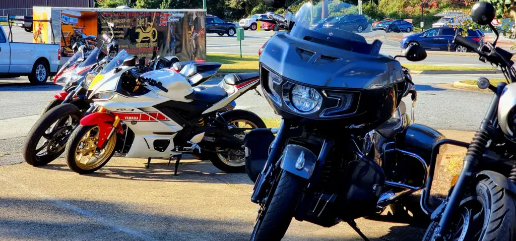 A large black motorcycle and a white sport bike are parked in a lot with cars and trees behind them.