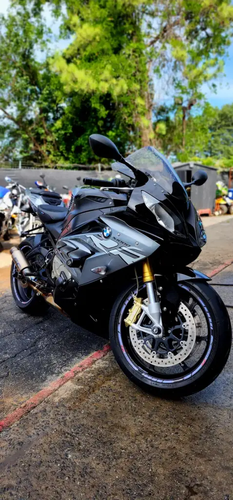 A black BMW sport motorcycle stands on wet pavement outdoors, with trees and other motorcycles behind it.