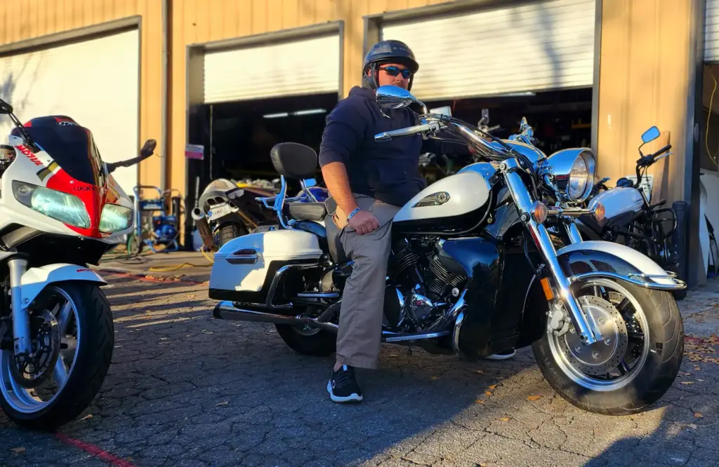 A man in a helmet and sunglasses sits on a white motorcycle outside a garage with other bikes nearby.