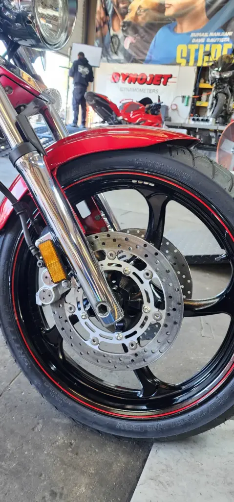 A close-up shows a motorcycle front wheel with disc brake in a workshop, mechanics and tools behind.