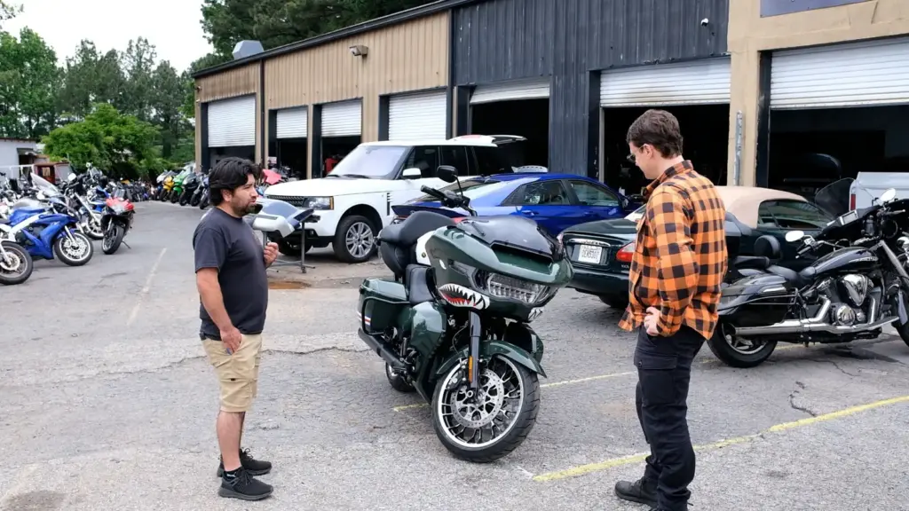 Two men stand by a custom motorcycle with a shark mouth design outside a garage with other vehicles.