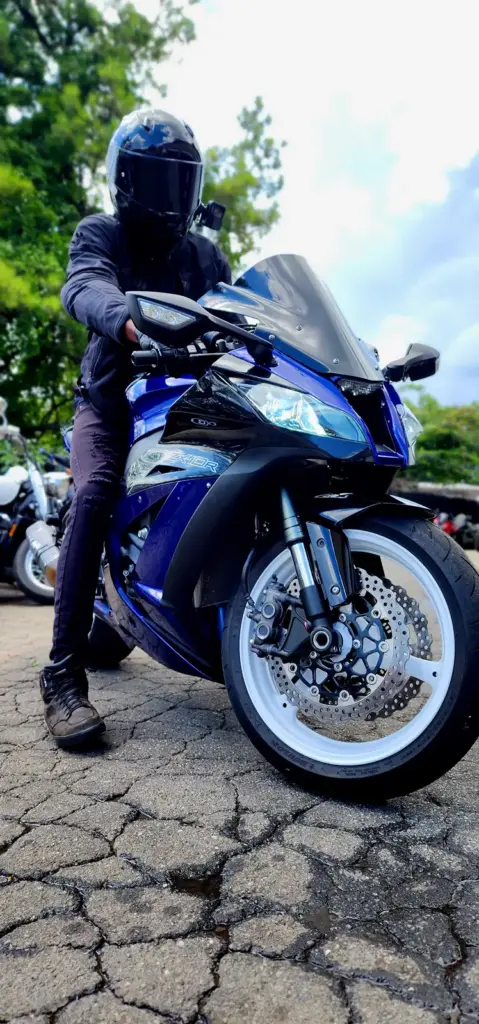 Person in black riding gear sits on a blue sport motorcycle parked on cracked outdoor pavement.